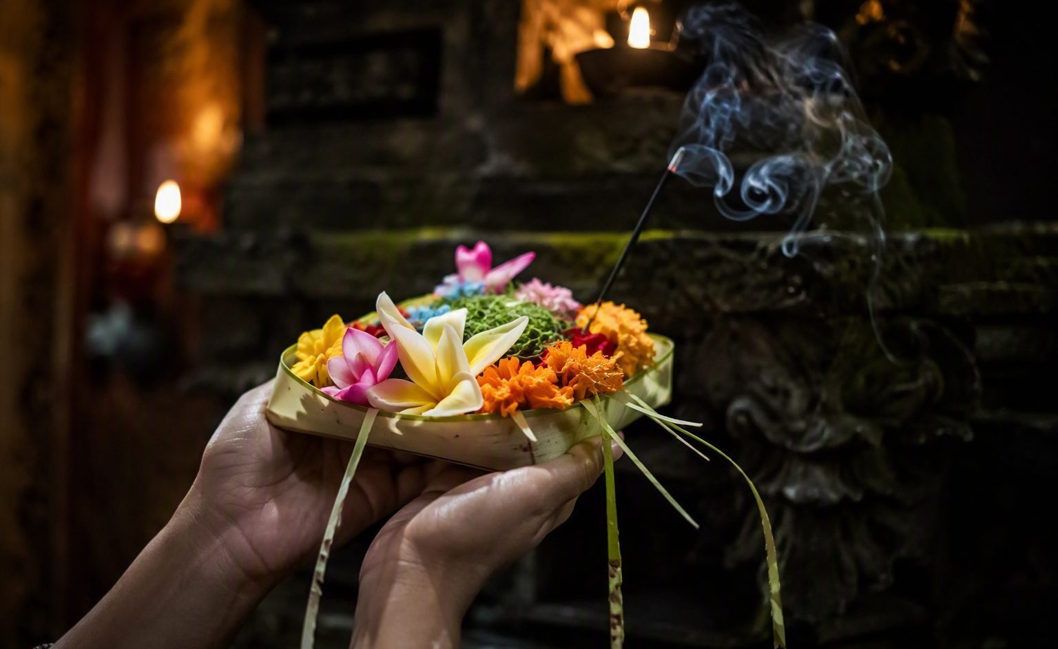 Hands offering a canang sari of flowers and incense